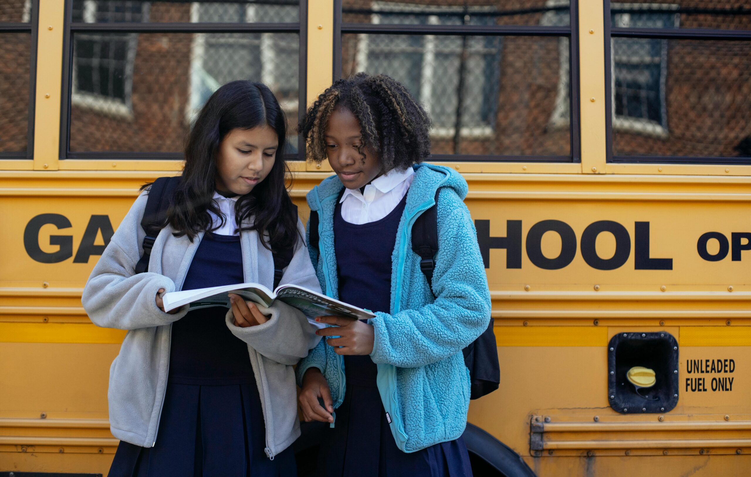 Multiethnic schoolchildren in uniforms watching exercise book near yellow school bus reflecting building in town